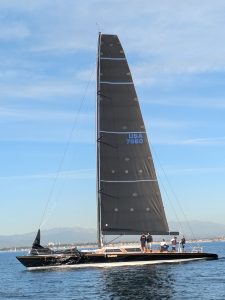 Ragtime slicing through calm Pacific waters off the Southern California coast, with her sleek black hull and tall carbon rig catching the morning light. The crew stands poised, focused, and ready, as the legendary racing yacht glides effortlessly beneath a clear blue sky.