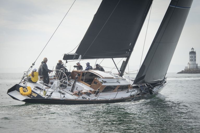Ragtime under sail near the breakwater in Long Beach, California. With her bold sail marked USA 7960 and crew preparing at the mast, the yacht’s striking silhouette stands out against palm trees and a rugged shoreline—where modern grit meets timeless elegance.